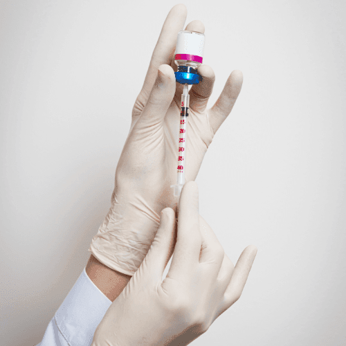 Gloved hands holding a syringe and vial, preparing a needle for injection with a white background.